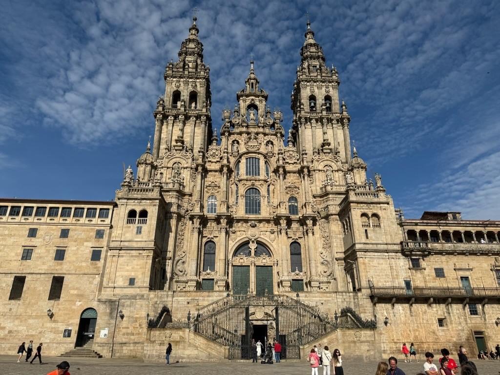 The Santiago de Compostela Cathedral, a giant light-brown structure with many architectural features such as round arches and windows, is shown with the blue sky in the background.