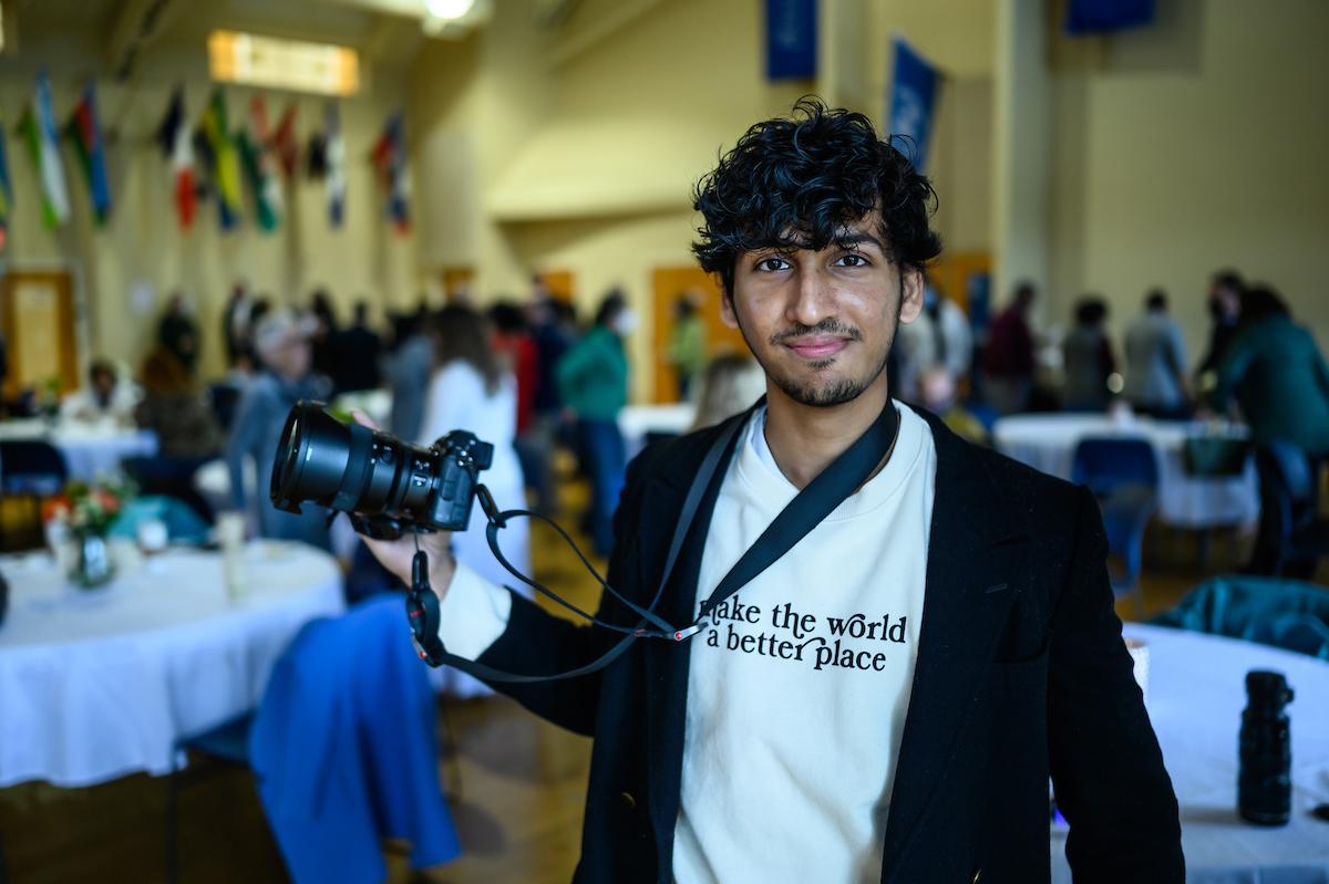 Male student holding a camera wearing a shirt that says A Chance to Make the World a Better Place